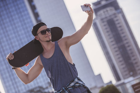 Stylish guy in casual clothes, cap and glasses is holding a skateboard and smiling while doing selfie using a smart phoneの写真素材