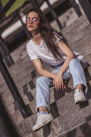 Stylish dark-haired girl in casual clothes and glasses is enjoying the sun while sitting on stairs outdoorsの写真素材