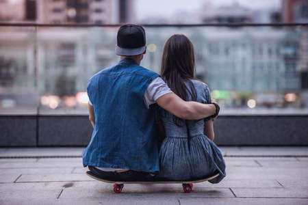 Back view of stylish young couple hugging while sitting on the skateboard and enjoying the viewの写真素材
