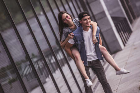 Stylish young couple is having fun and smiling while walking outdoors, girl is sitting pickabackの写真素材