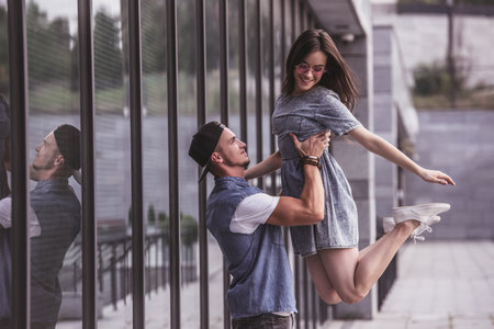 Stylish young couple is having fun and smiling while walking outdoors, guy is lifting his girlfriend