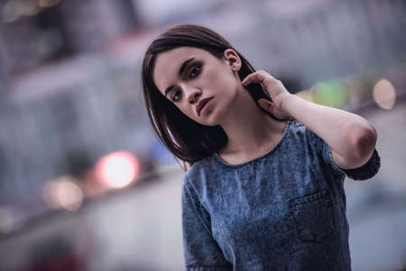 Portrait of beautiful dark-haired girl in jean dress looking at camera, on the city backgroundの写真素材