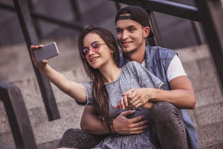 Stylish young couple is doing selfie using a phone and smiling while sitting on the skateboard on stairsの写真素材