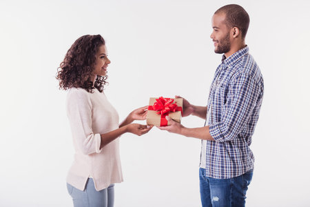 Beautiful Afro American girl and handsome guy are holding a present, looking at each other and smiling, isolated on whiteの写真素材