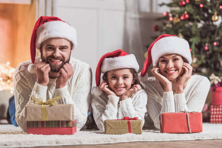Merry Christmas and Happy New Year! Beautiful mom, dad and daughter in Santa hats are looking at camera and smiling while lying near Christmas presentsの写真素材