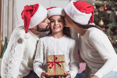 Merry Christmas and Happy New Year! Cute little girl is holding a present, looking at camera and smiling while her parents are kissing herの写真素材
