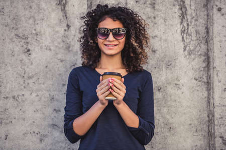 Attractive girl in casual clothes and sun glasses is holding a cup of coffee, looking at camera and smiling, on concrete wall backgroundの写真素材