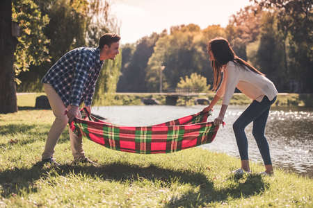 Beautiful young couple is putting a plaid on the grass and smiling while resting in the parkの写真素材