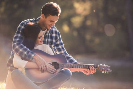 Beautiful young couple is resting on a plaid in the park, guy is teaching his girlfriend how to play guitarの写真素材