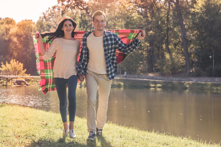 Beautiful young couple holding hands and a plaid, and smiling while walking in the parkの写真素材