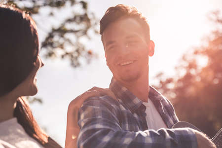 Beautiful young couple is singing and smiling while resting on a plaid in the park, guy is playing guitarの写真素材