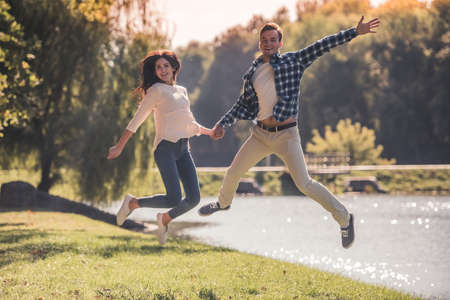 Beautiful young couple is holding hands, jumping at camera and smiling while walking in the parkの写真素材