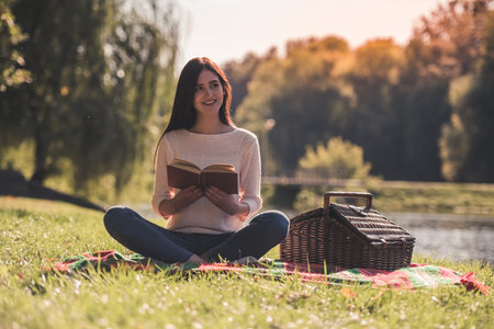 Beautiful young girl is reading a book and smiling while resting on a plaid in the parkの写真素材