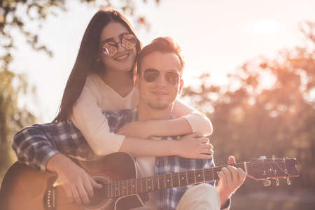 Beautiful young couple is hugging and smiling while resting in the park, guy is playing guitarの写真素材
