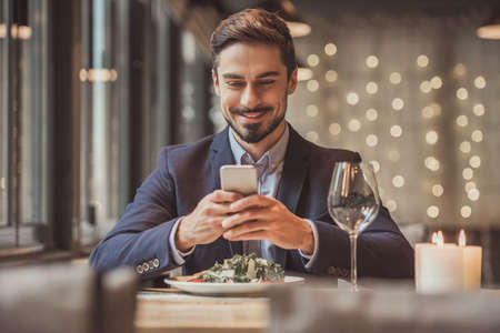 Handsome businessman in suit is using a smart phone and smiling while sitting in restaurantの写真素材