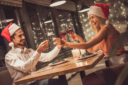 Beautiful young couple in Santa hats is looking at each other, clinking glasses of wine together and smiling while celebrating New Year in a restaurantの写真素材