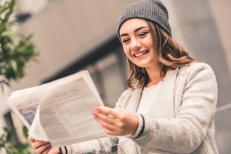 Beautiful girl in coat and cap is reading a newspaper and smiling while resting outdoorsの写真素材