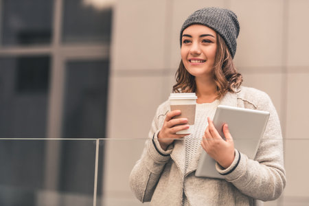 Beautiful girl in coat and cap is drinking coffee, holding a digital tablet and smiling while...