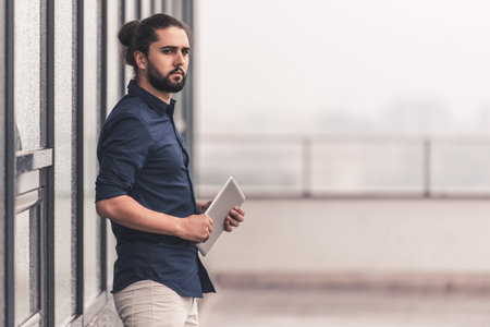Stylish young businessman with long dark hair is holding a digital tablet while standing outdoors