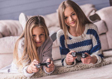 Two attractive teenage girls in casual clothes are playing video game console and smiling while...
