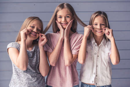 Three attractive teenage girls in casual clothes are looking at camera and smiling, two are making mustaches of their friend's hair, on gray wall backgroundの写真素材