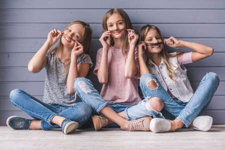 Three attractive teenage girls in casual clothes are making moustaches of their hair at camera while sitting on the floor, on gray wall backgroundの写真素材