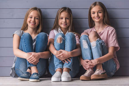 Three attractive teenage girls in casual clothes are looking at camera and smiling while sitting on the floor, on gray wall backgroundの写真素材