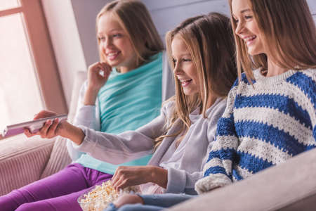 Three attractive teenage girls in casual clothes are eating popcorn and smiling while watching TV on couch at homeの写真素材