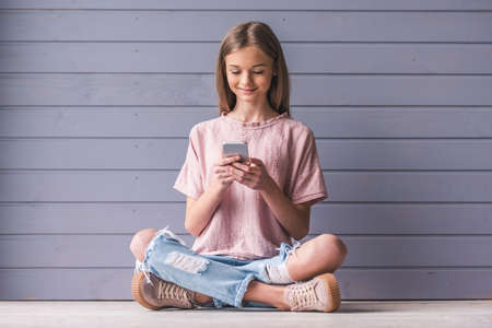 Attractive teenage girl in casual clothes is using a smartphone and smiling while sitting on the floor, on gray wall backgroundの写真素材