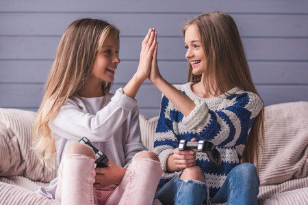 Two attractive teenage girls in casual clothes are playing video game console, giving high five and...