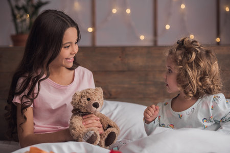 Cute little girls are talking and smiling while sitting on bed at home on Christmas