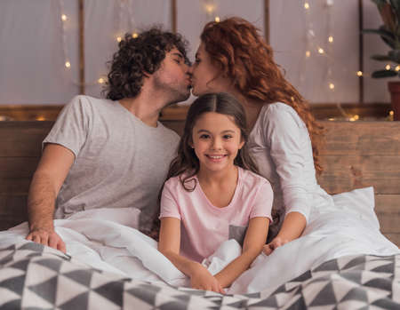 It's Christmas time! Cute little girl and her parents are smiling while lying in bed at home, parents are kissingの写真素材