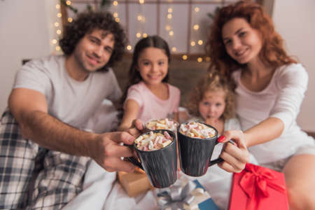 It's Christmas time! Happy parents and their daughters are holding cups and smiling while sitting on bed with presentsの写真素材