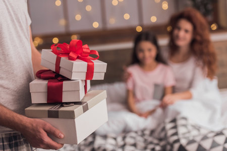 It's Christmas time! Cute little girl and her beautiful mom are lying in bed, dad is holding presents in the foregroundの写真素材