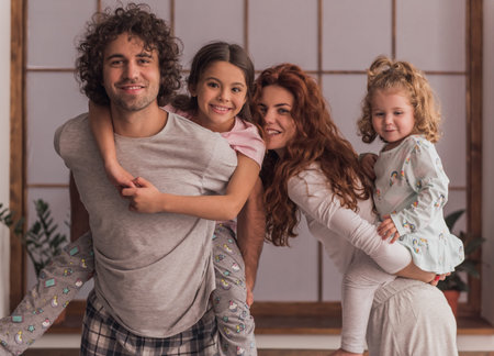 Happy parents and their cute daughters are smiling while spending time together at home, girls are...
