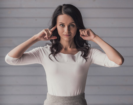 Attractive young woman is covering her ears and looking at camera, on gray background