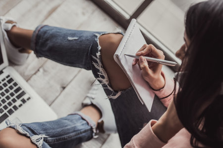 Attractive young woman in casual clothes is making notes, talking on the mobile phone and smiling while working with a laptop at homeの写真素材