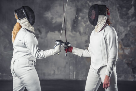 Female fencers in protective clothing are fighting on dark gray background