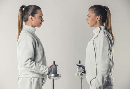 Beautiful female fencers in protective clothing are holding weapon and looking at each other, on gray background, side viewの写真素材