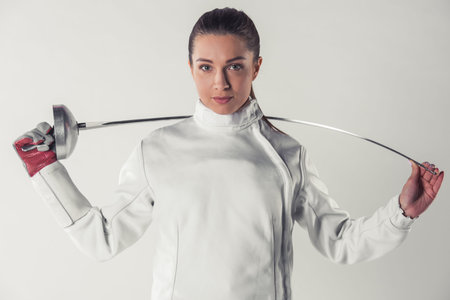 Beautiful female fencer in protective clothing is holding a weapon and looking at camera, on gray...