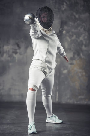 Female fencer in protective clothing is using weapon while practicing on dark gray background