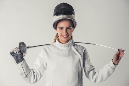 Attractive female fencer in protective clothing is holding a weapon, looking at camera and smiling,...