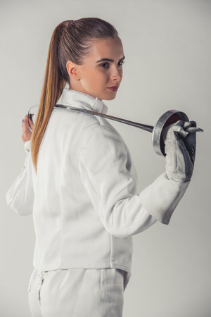 Attractive female fencer in protective clothing is holding a weapon, on gray background