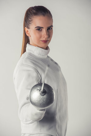 Attractive female fencer in protective clothing is holding a weapon and looking at camera, on gray...
