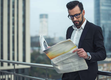 Handsome businessman in suit and glasses is reading newspaper while standing on the balconyの写真素材