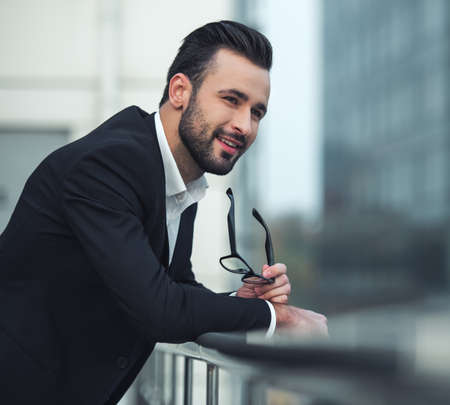 Handsome businessman in suit is holding glasses, looking away and smiling while leaning on the balconyの写真素材
