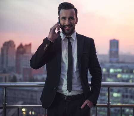 Handsome businessman in suit is talking on the mobile phone and smiling while standing on the balcony, beautiful sunset behind himの写真素材