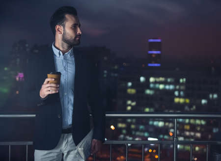 Handsome businessman in suit is holding a cup of coffee and looking away while standing on the balcony in the eveningの写真素材