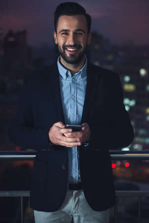 Handsome businessman in suit is holding a smart phone, looking at camera and smiling while standing on the balcony in the eveningの写真素材