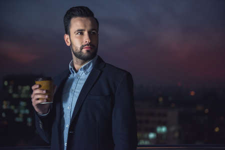 Handsome businessman in suit is holding a cup of coffee and looking away while standing on the balcony in the eveningの写真素材